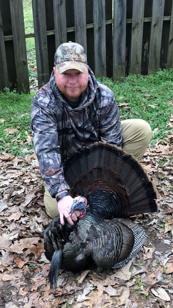 Man in camouflage kneeling next to a wild turkey, displaying the bird's fanned tail feathers. He is smiling. Outdoors with wooden fence in the background.