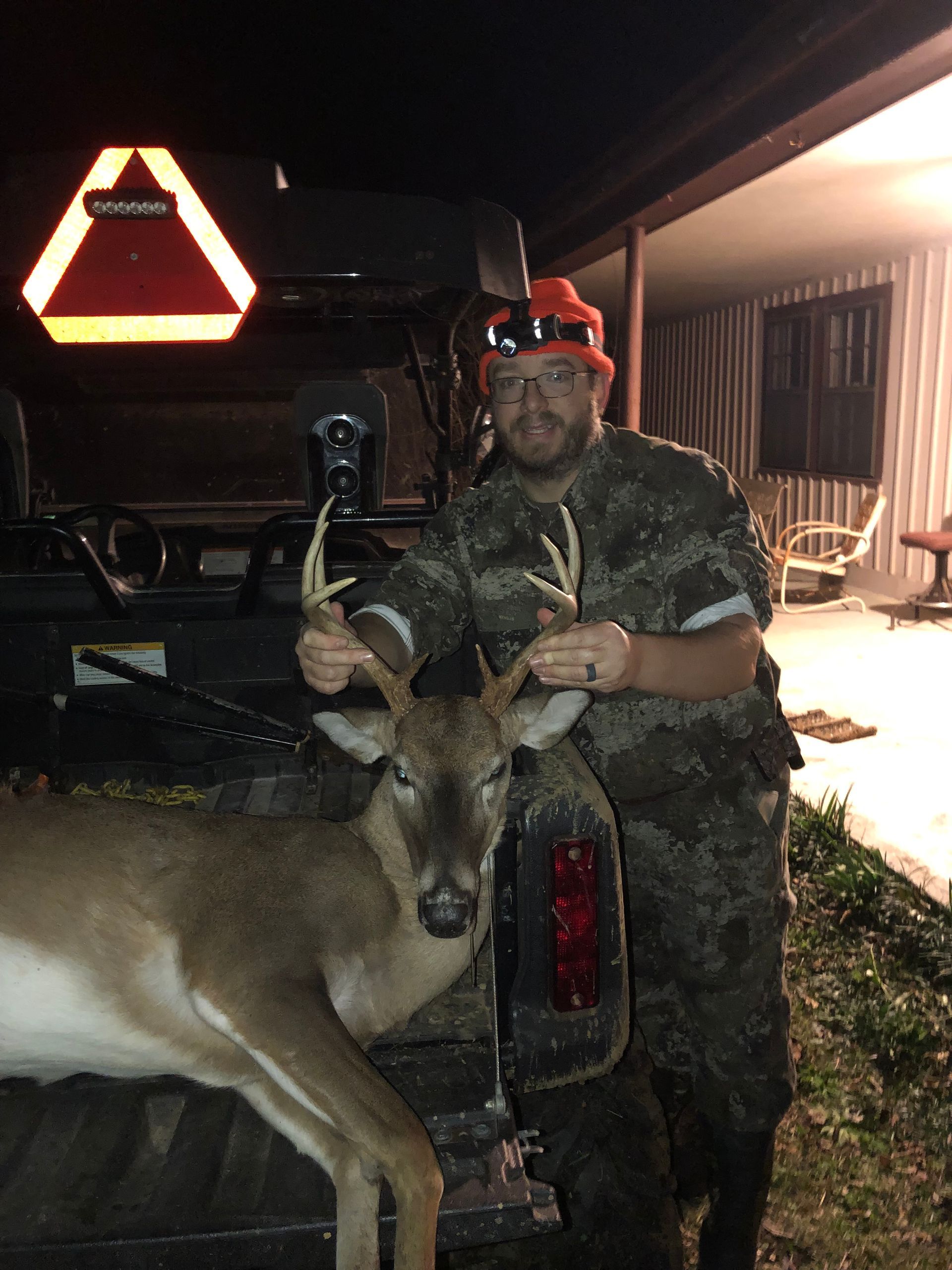 A hunter in camouflage holds a buck he shot. He's smiling, standing beside a utility vehicle at night.