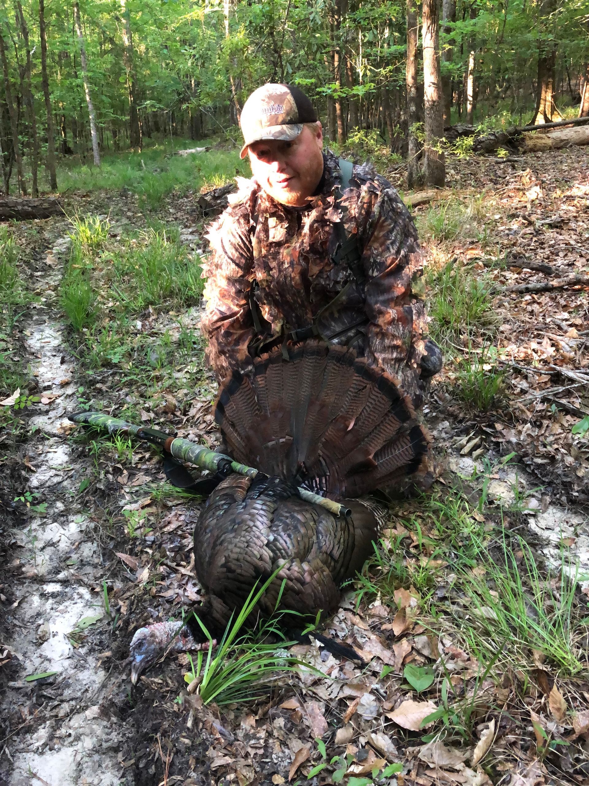 A man in camouflage kneels next to a harvested wild turkey in a wooded area. He smiles, holding the bird's fanned tail.