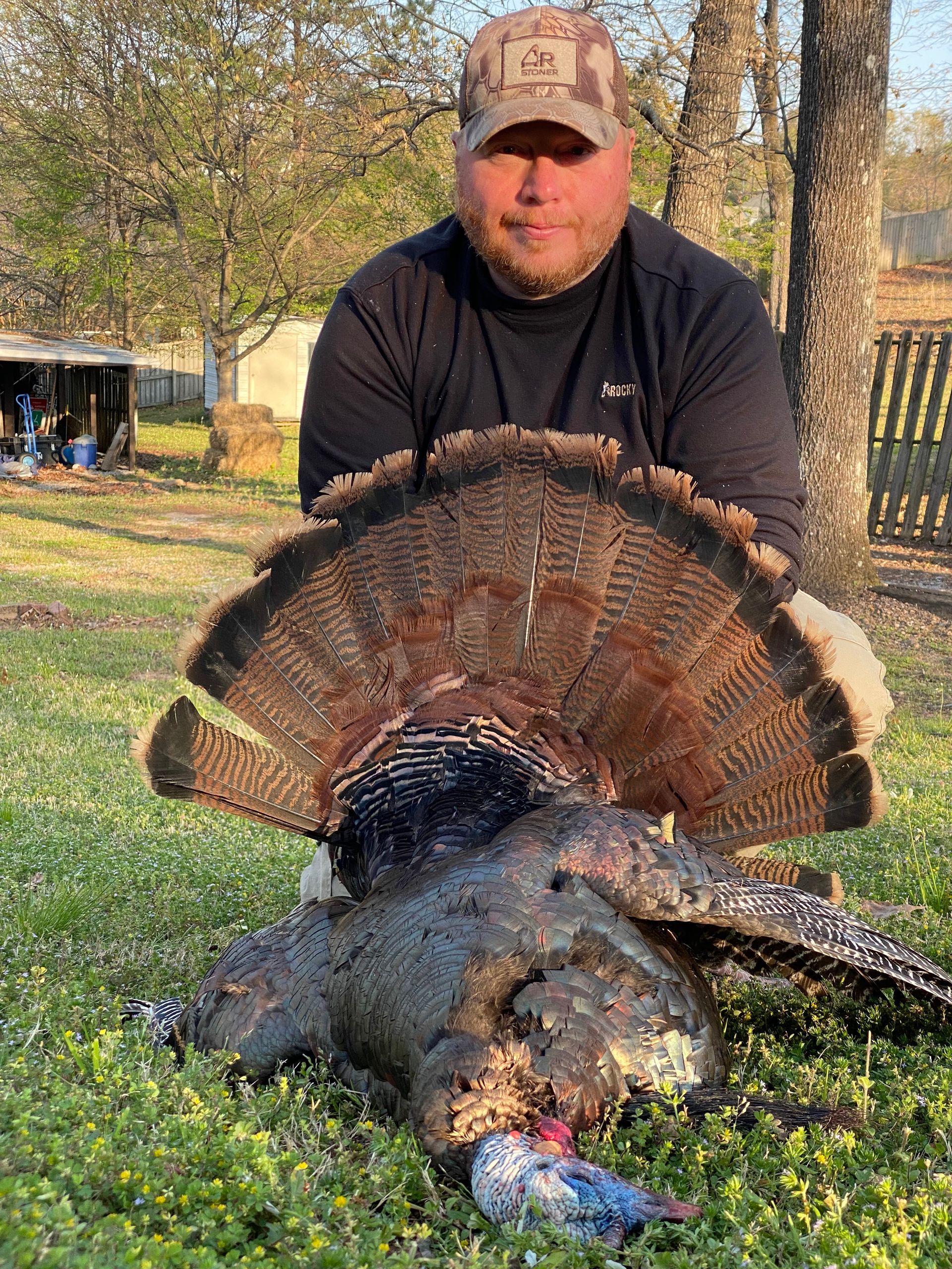 Man in a cap holds up a dead turkey with its feathers fanned out on green grass, outdoors.