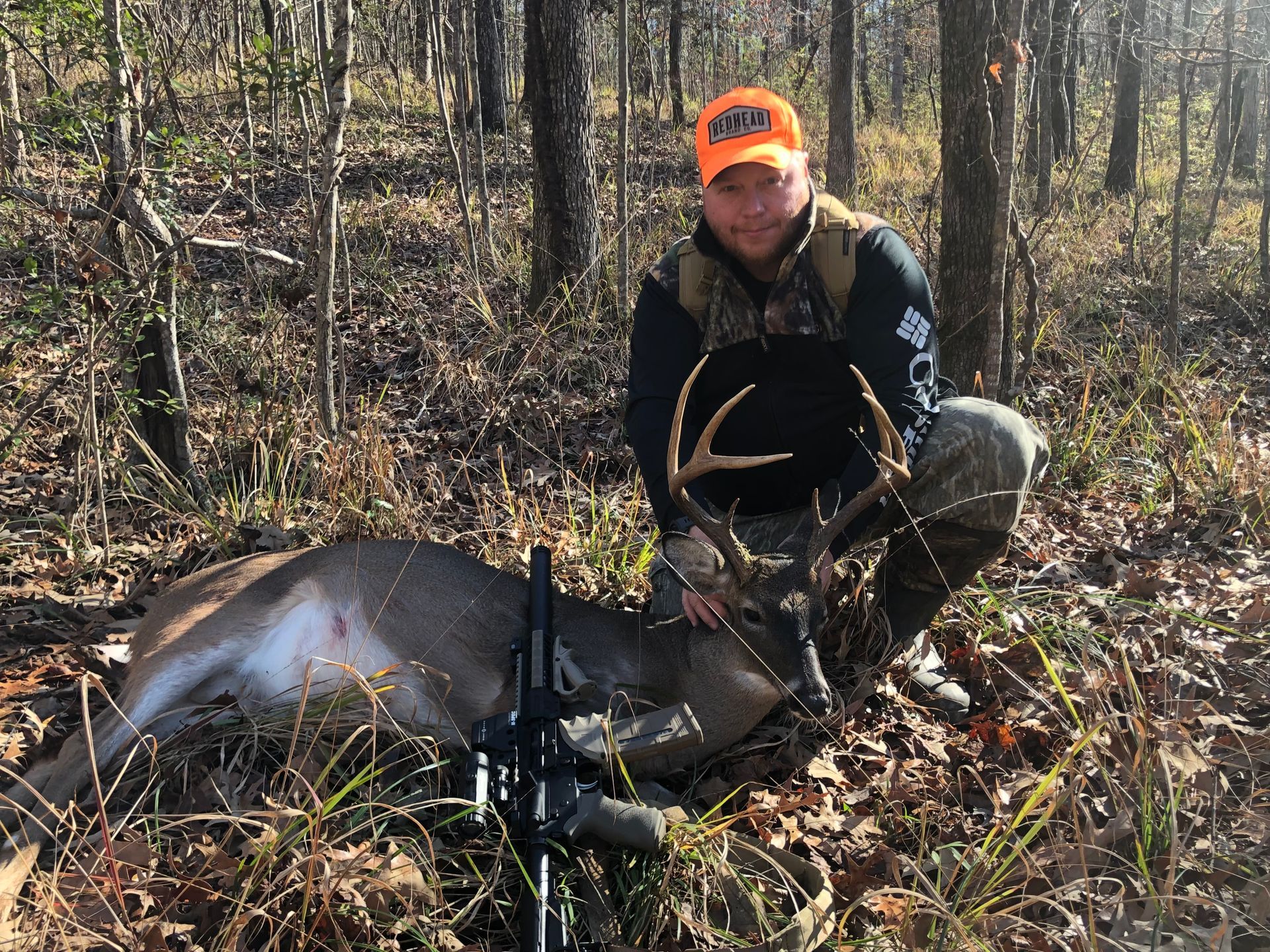 A man kneels beside a large buck in a forest. He wears hunting gear and smiles, and a rifle lies nearby.