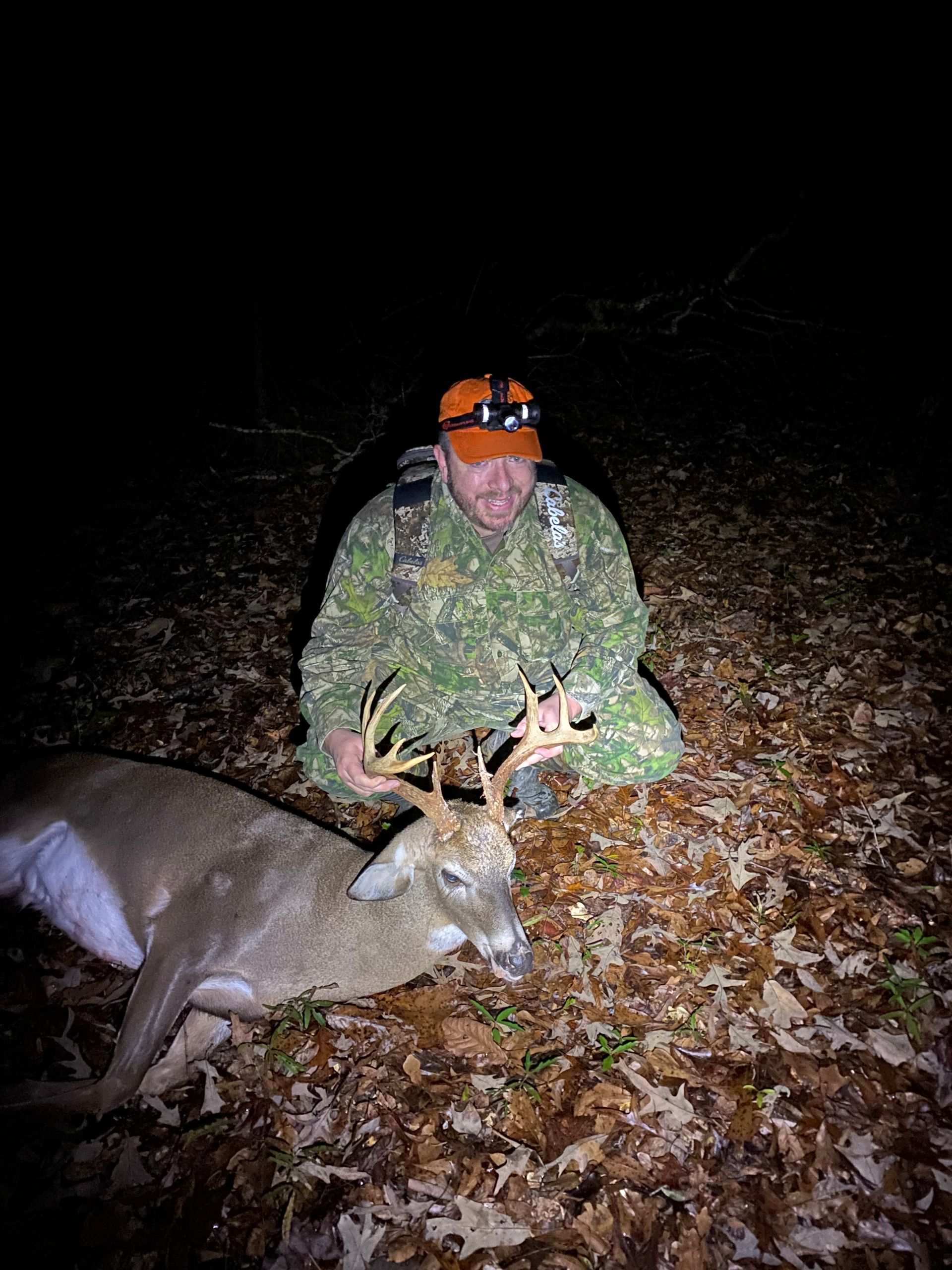 A man in camouflage kneels next to a large, dead buck with impressive antlers in a dark forest setting. He has an excited expression.