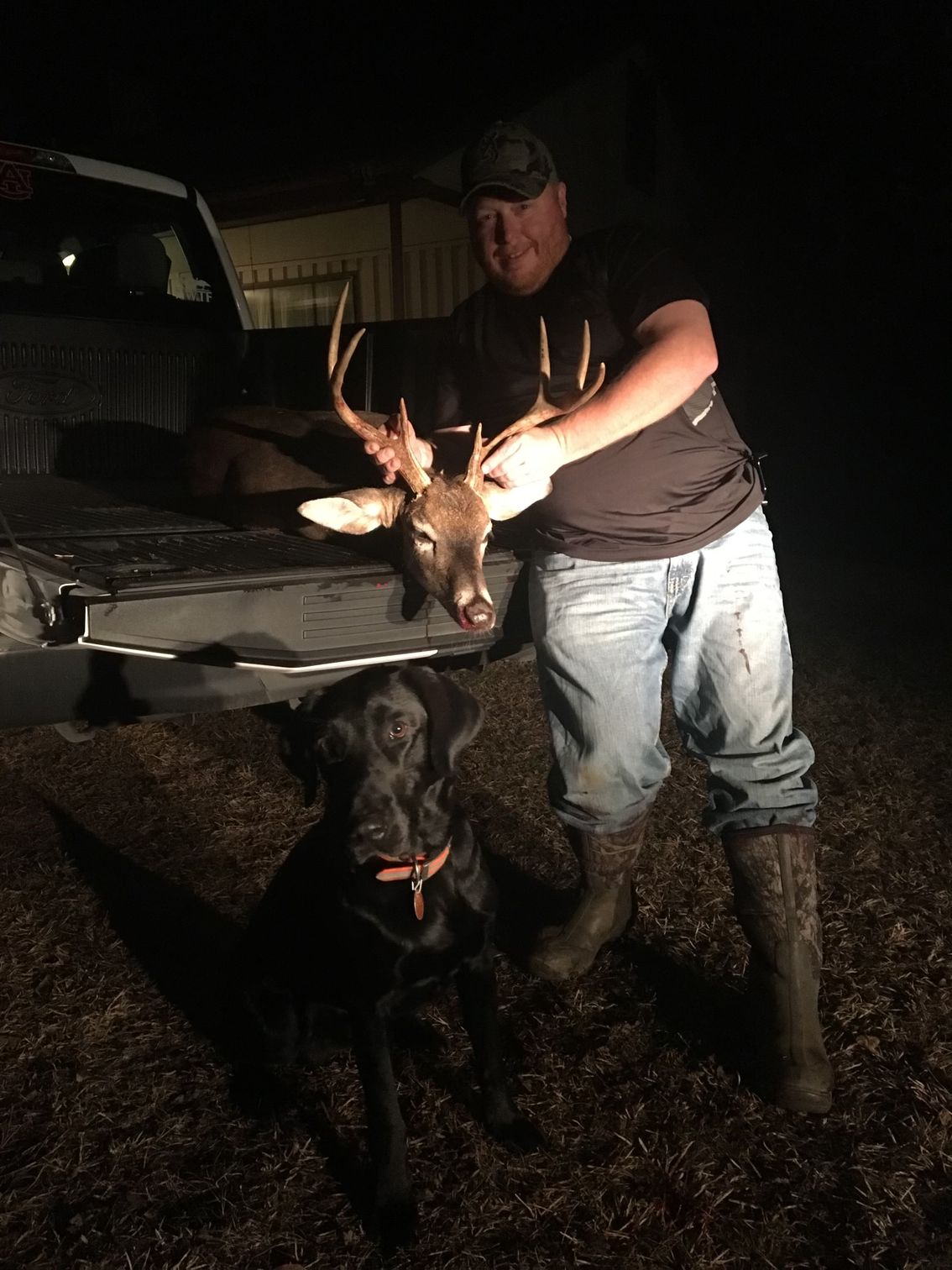 A man holding a deer head, standing next to a black dog and a pickup truck at night.