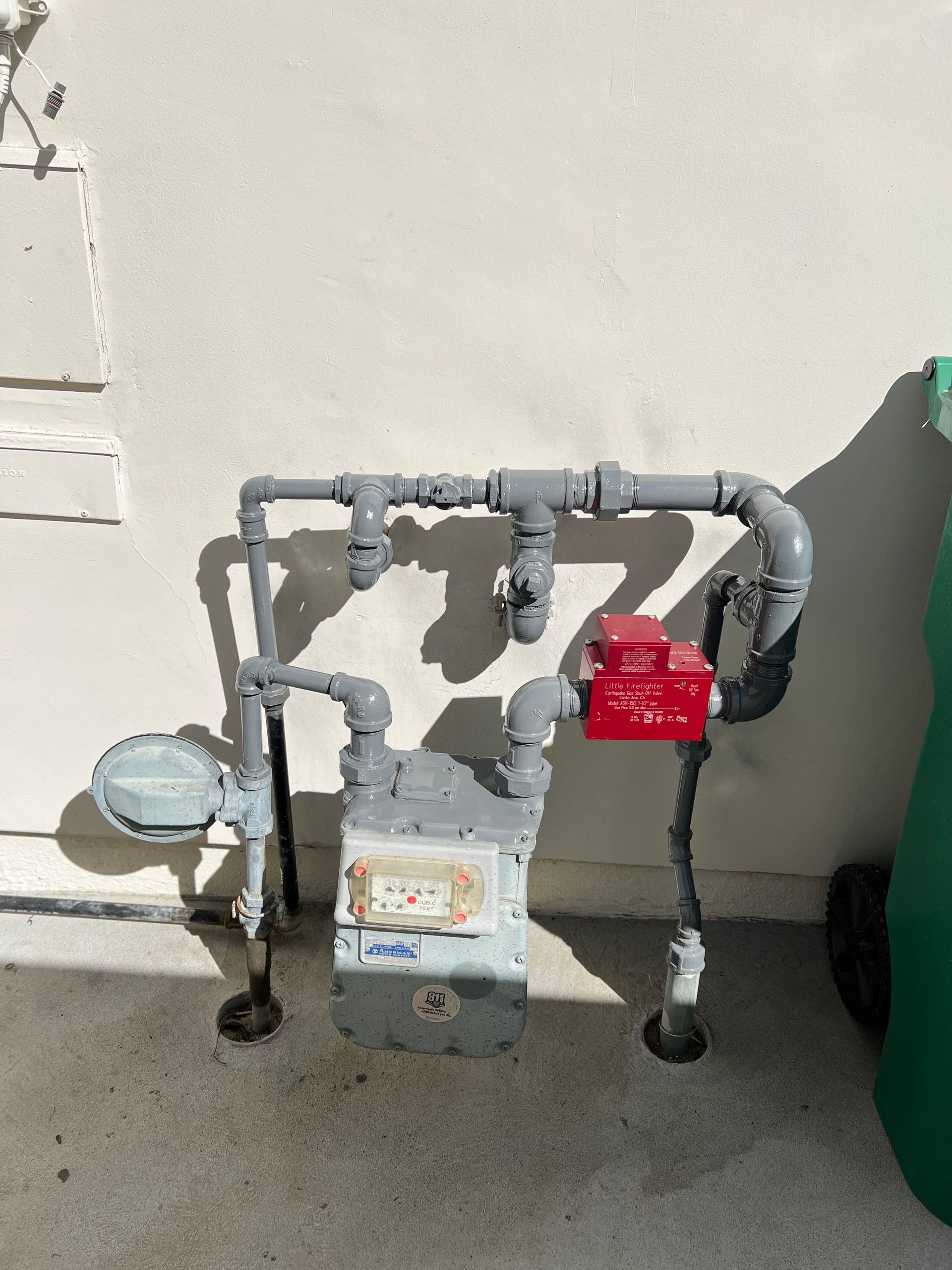 A gas meter is sitting next to a green trash can
