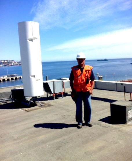 Un hombre con un chaleco naranja y un casco de seguridad se encuentra en un tejado con vistas al océano.