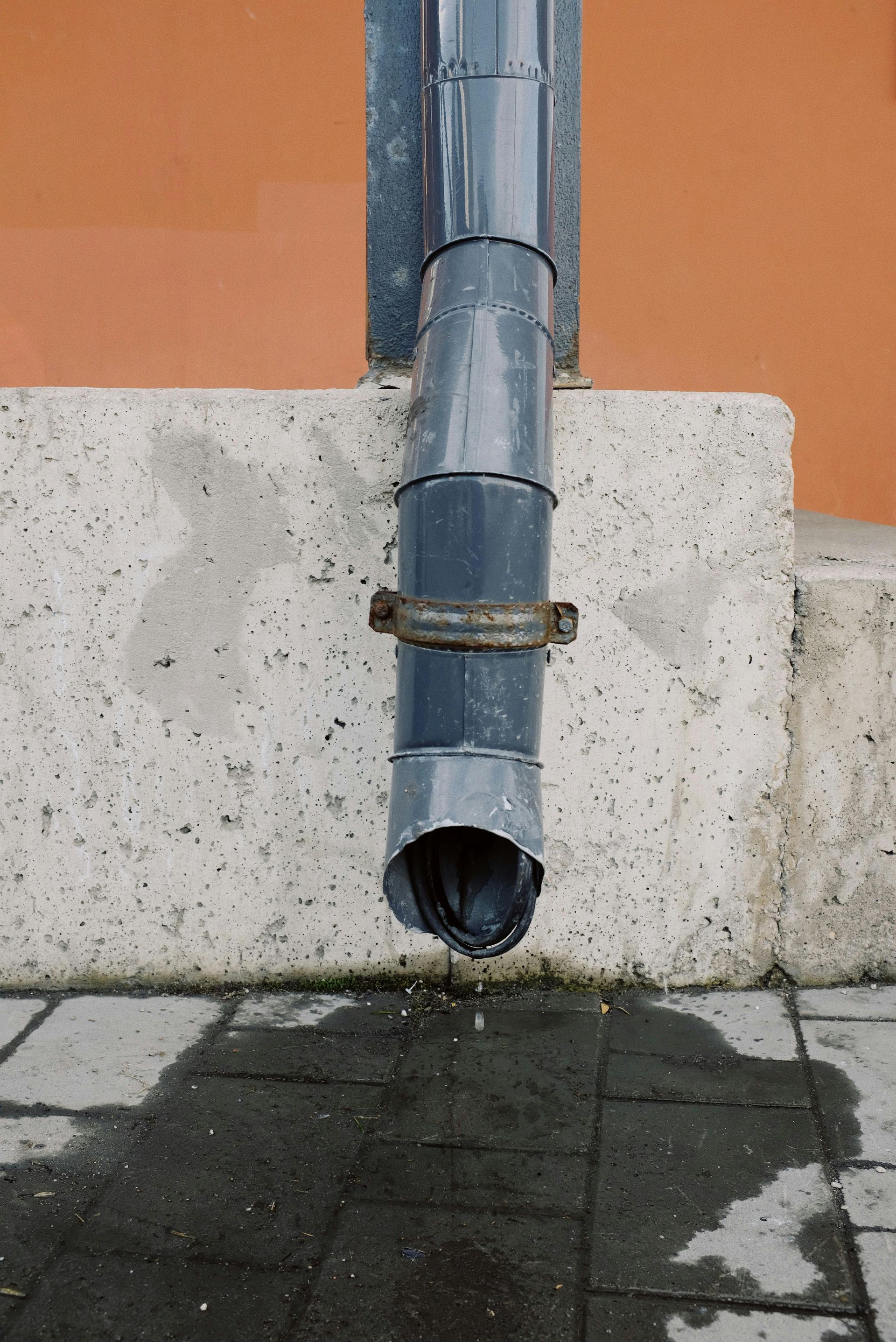 Rainwater draining from a gray downspout onto wet pavement, against an orange wall and concrete block.