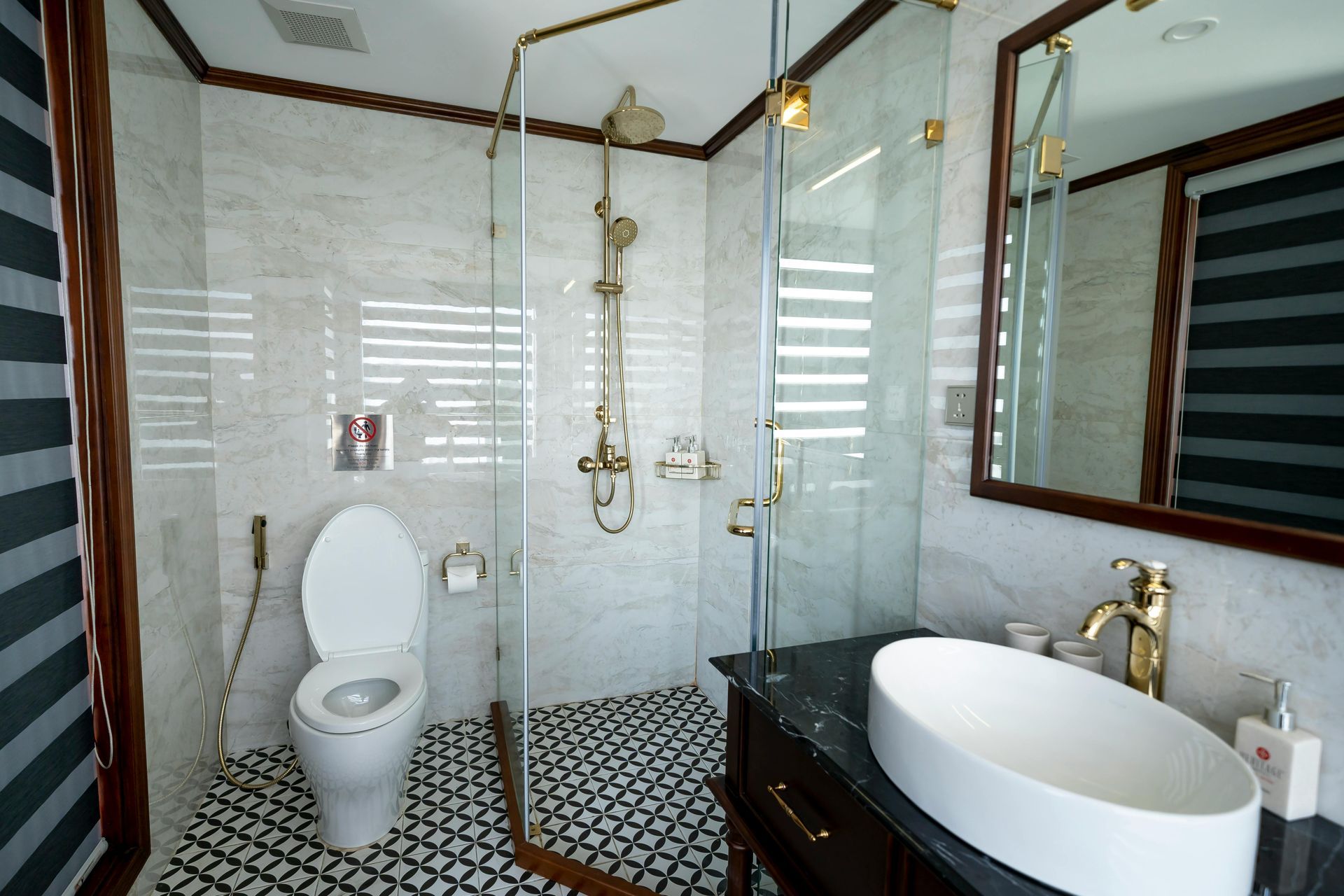 Bathroom with marble walls, glass shower, black and white floor tiles, and gold fixtures.