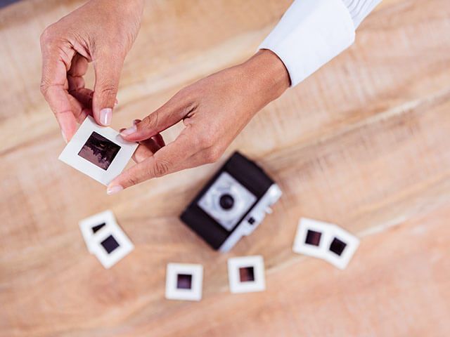 Hands holding a photo slide, with a small camera and other slides on a wooden surface.