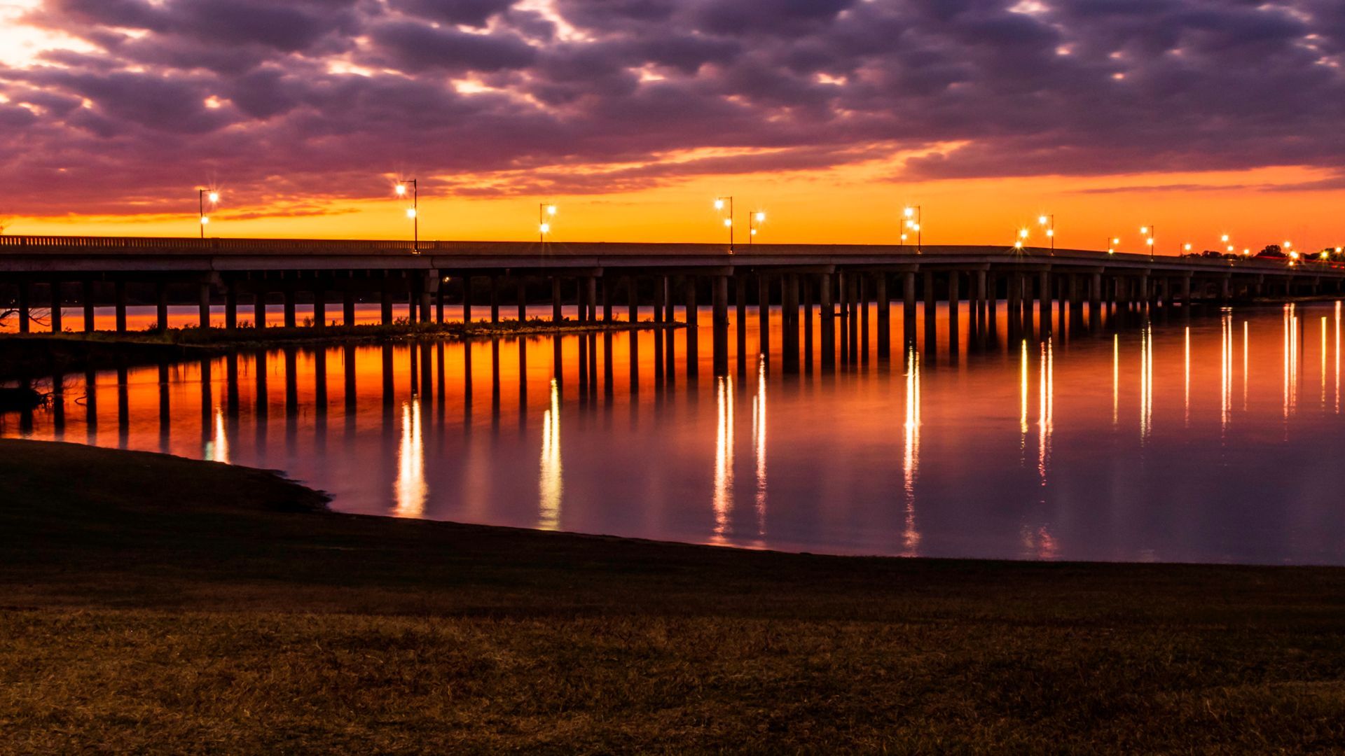 A bridge over a body of water at sunset with lights reflected in the water.