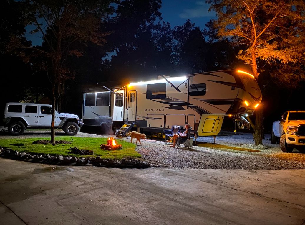 A group of people are sitting around a campfire in front of a rv at night.