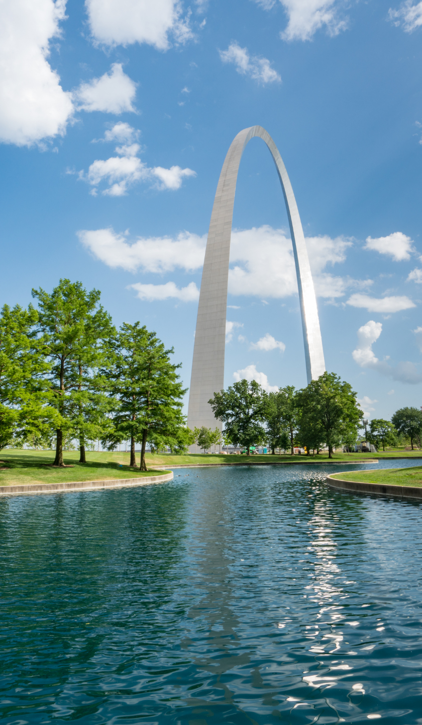 The Gateway Arch towers over a reflective pond with green trees under a bright blue sky with white clouds.