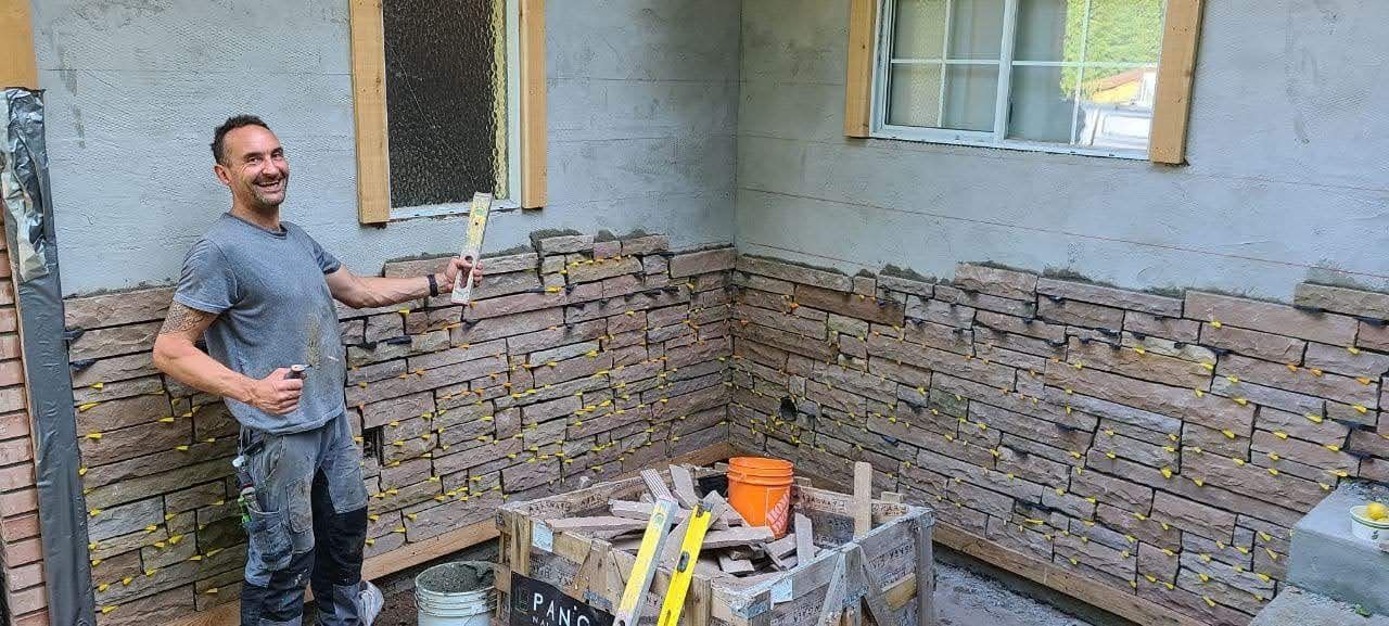 Chris Armbruster owner of Bricked Masonry is standing in front of a partly finished cultured stone veneer wall.