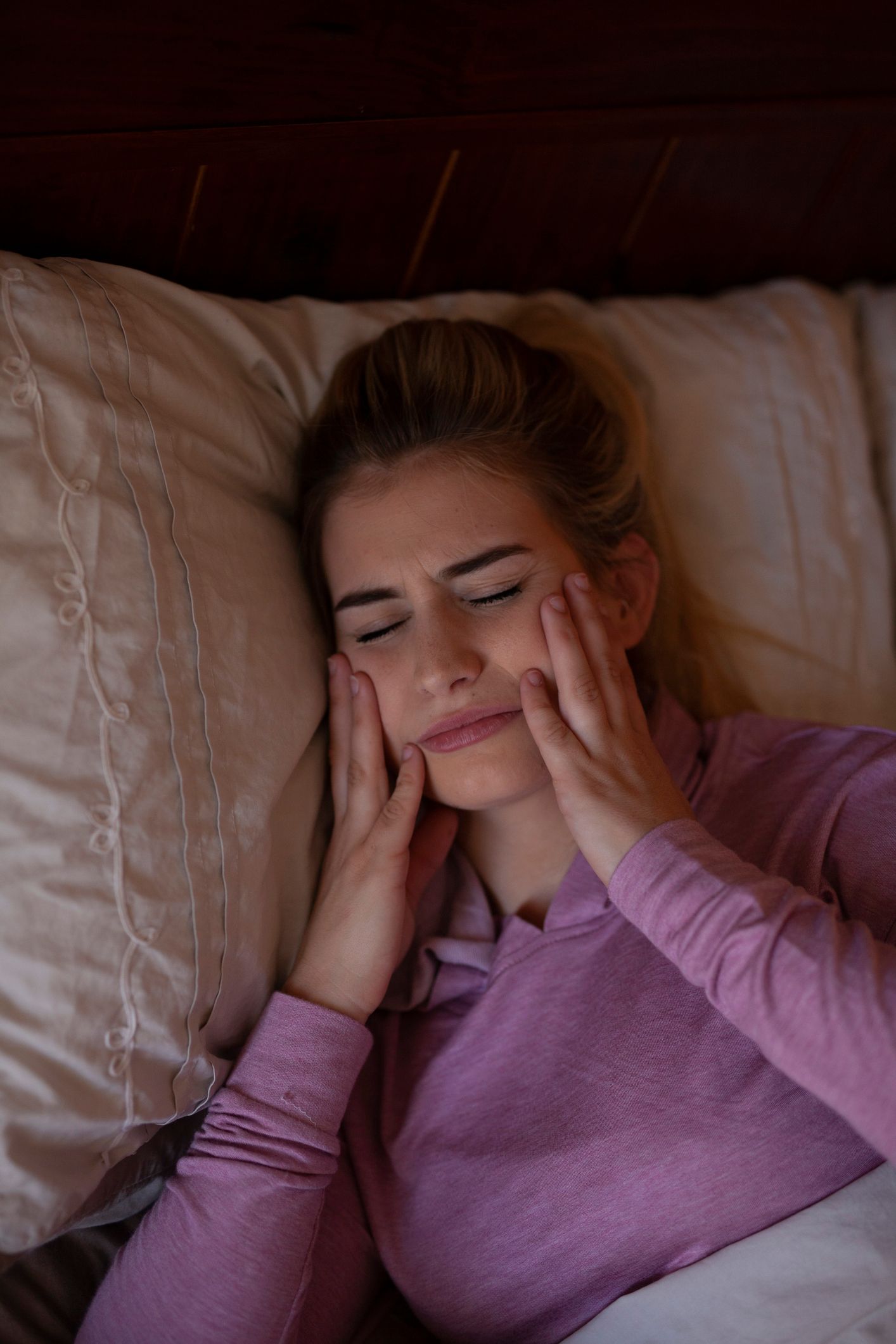 Woman in bed with hands on cheeks, appears to be in pain or discomfort, soft lighting.