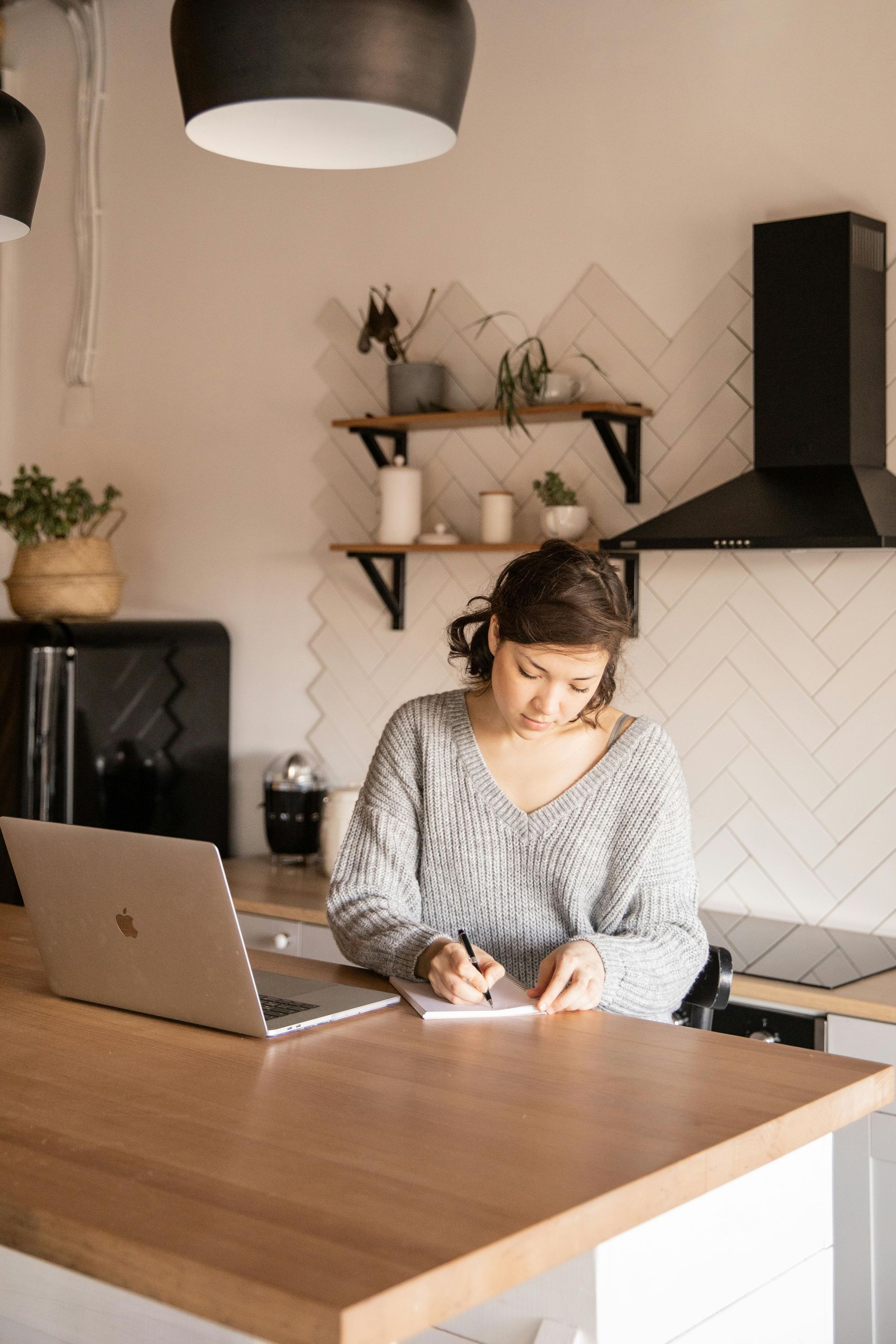 A woman is sitting at a kitchen counter with a laptop and writing in a notebook.