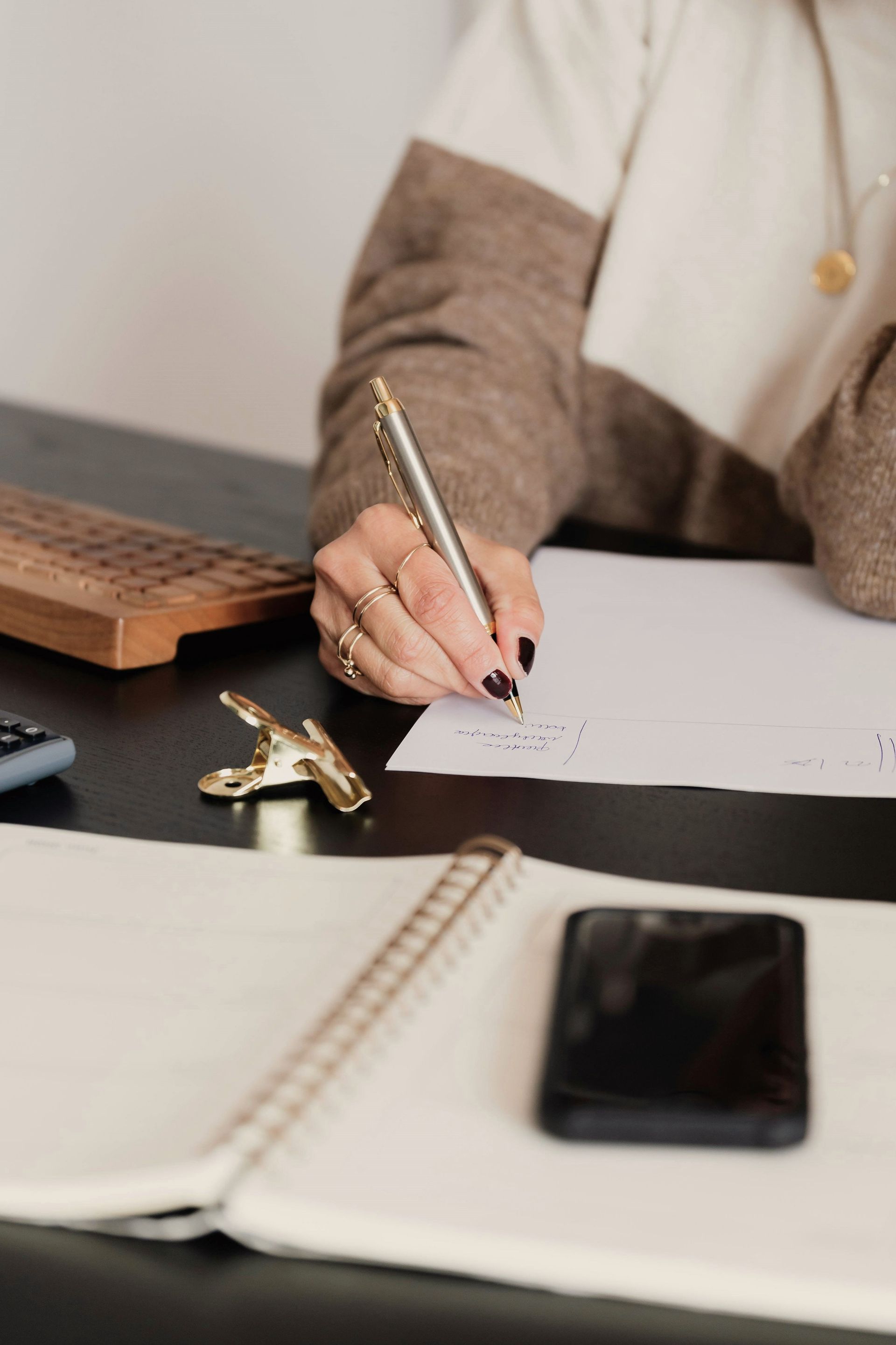 A woman is sitting at a desk writing in a notebook with a pen.