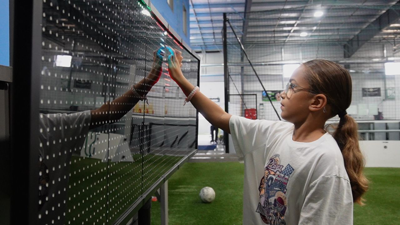 A young girl is standing in front of a wall with a soccer ball in the background.