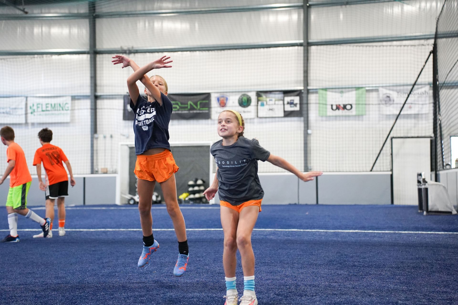 Two young girls are jumping in the air on a soccer field.