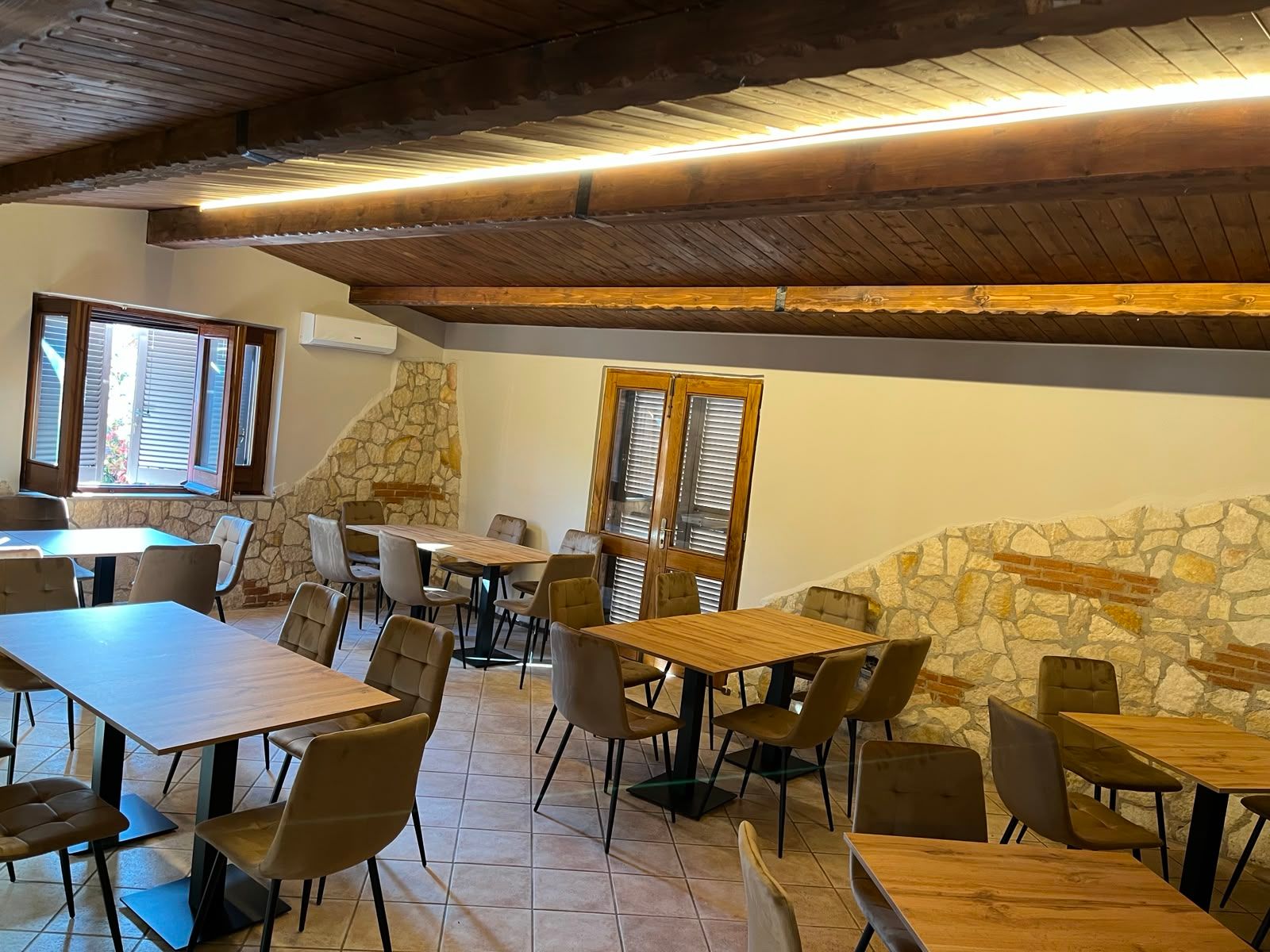 Accogliente caffè in stile rustico con travi in legno, pareti in pietra e tavoli e sedie vuoti vicino a una finestra.