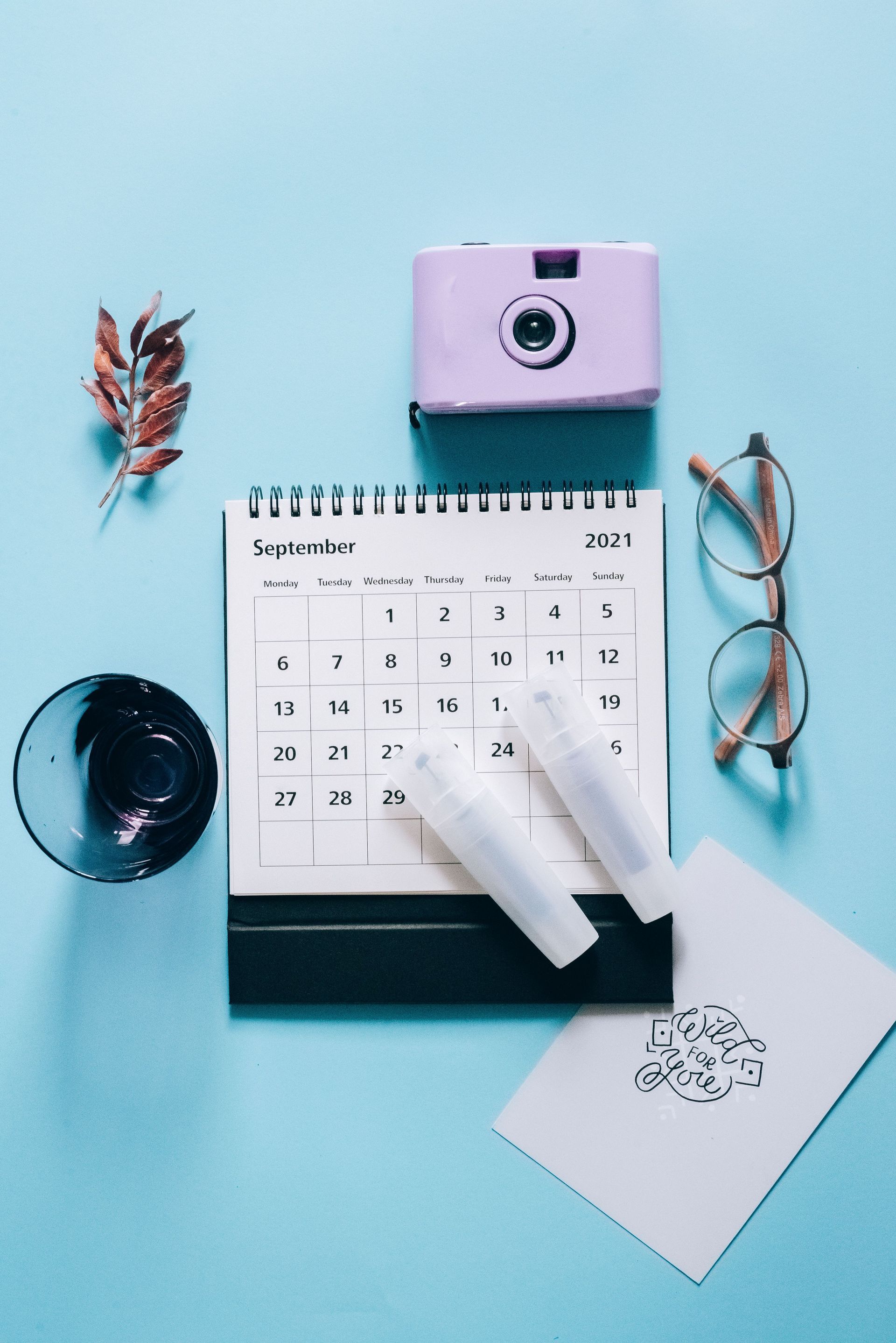 Flat lay of a blue desk with calendar, pink camera, glasses, candle, and stationery on a teal background