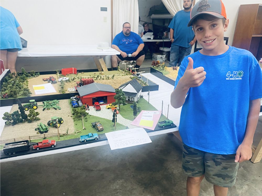 Boy in blue shirt giving a thumbs-up beside a model display table at an indoor exhibit.