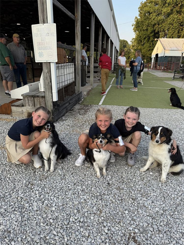 Three smiling people kneel with four dogs on gravel outside a barn at dusk.