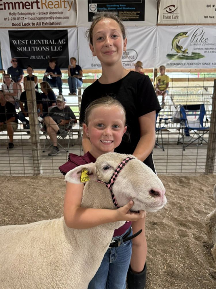 Two smiling girls with a white sheep at a livestock show under a canopy