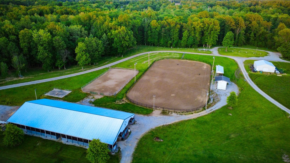 Aerial view of a rural sports field with a dirt infield, blue-roofed building, and surrounding forest.