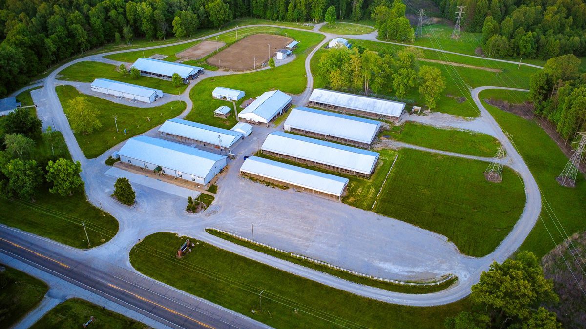 Aerial view of an industrial facility with white-roofed buildings, paved lots, and surrounding green trees.