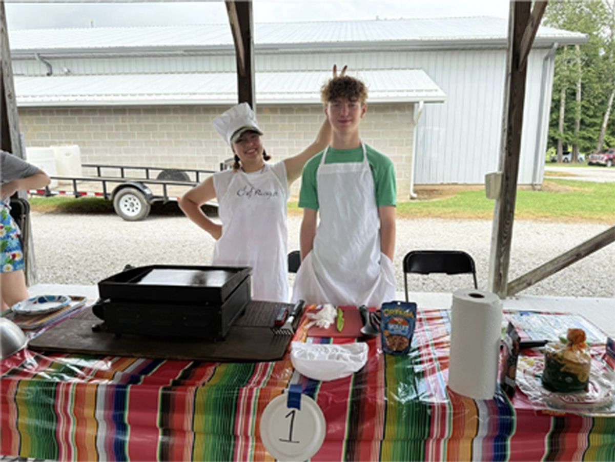 Two people behind a food table at an outdoor pavilion, with dishes and a striped tablecloth.