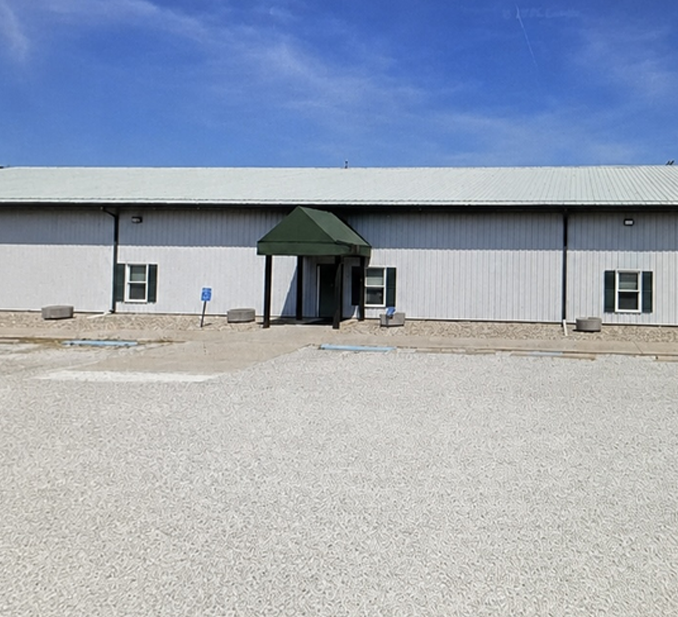 Plain building entrance with green awning, white metal siding, and gravel lot under a blue sky