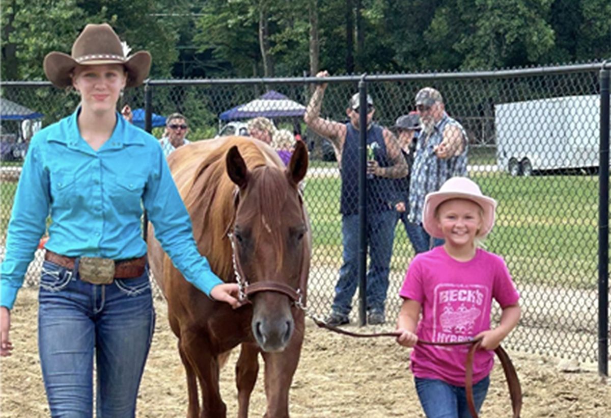 Woman and child leading a pony at an outdoor fair, with spectators and a fence in the background