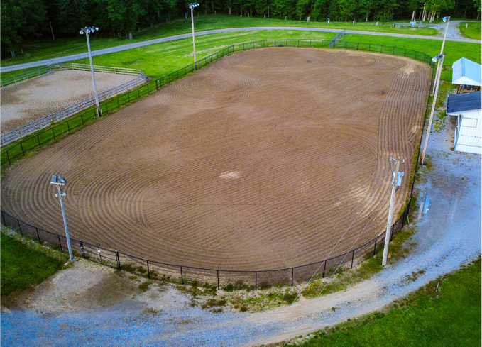 Empty dirt riding arena bordered by a fence and lights, surrounded by green grass and a dirt path