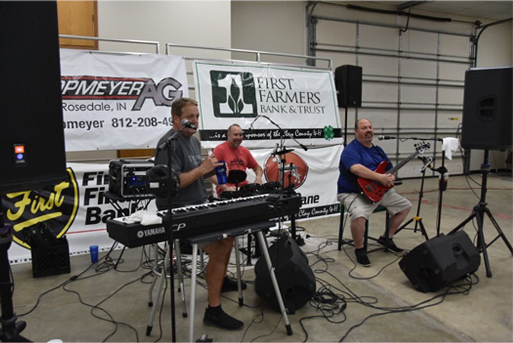 Three musicians performing indoors with keyboards, bass, and microphone stands at a local event