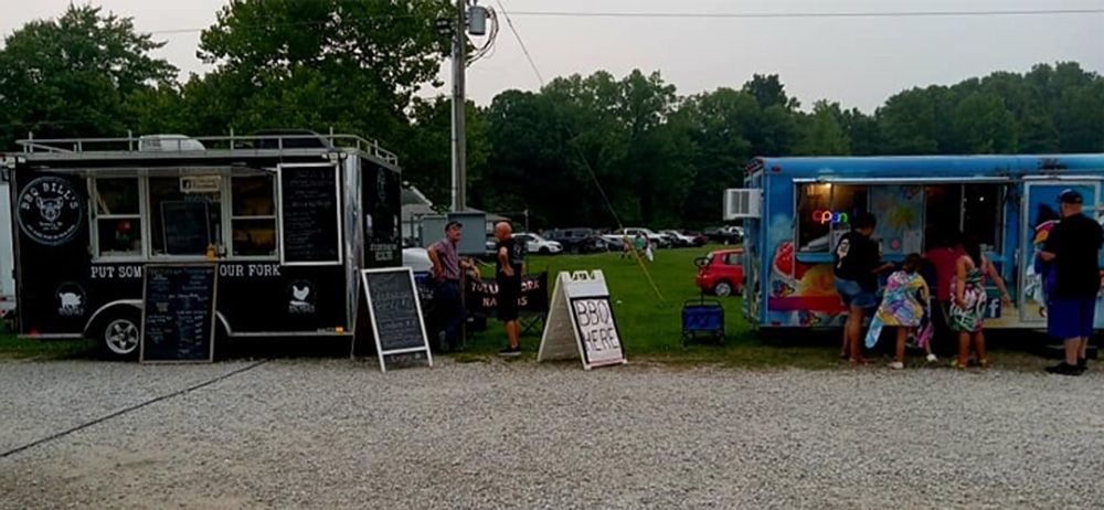 Two food trucks at an outdoor market, with people browsing beside a blue truck and a black truck.