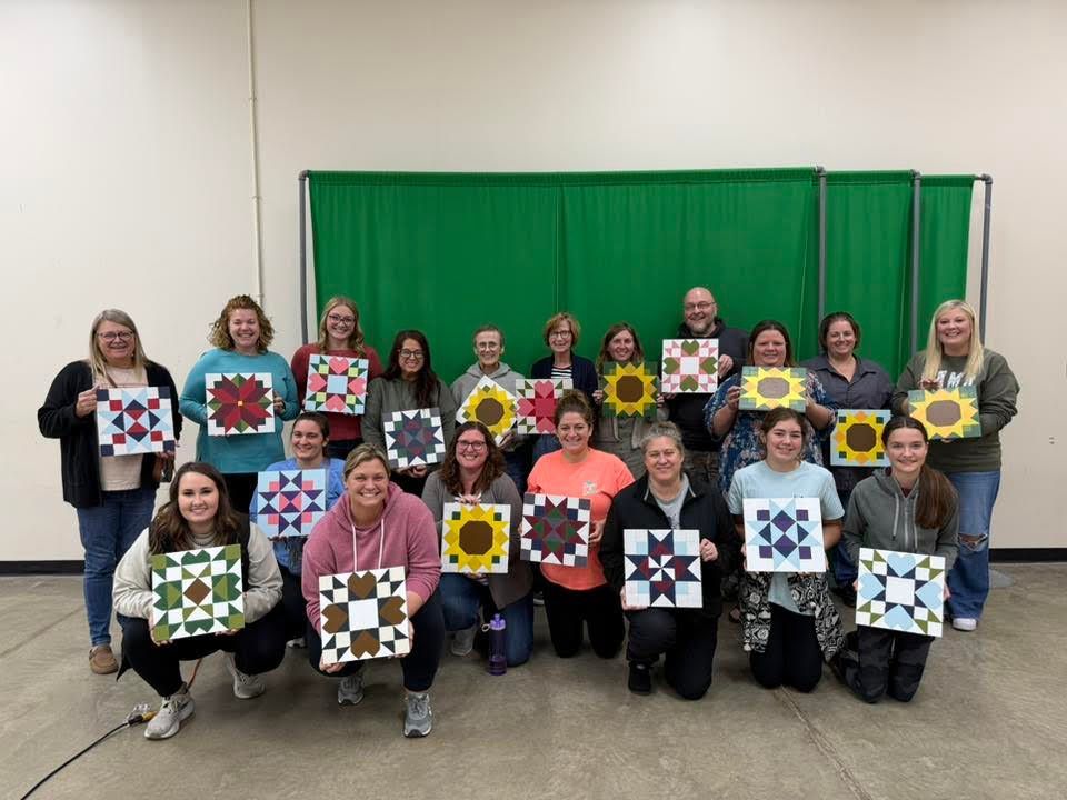 Group of people posing indoors, each holding a colorful patchwork quilt square in front of green curtains.