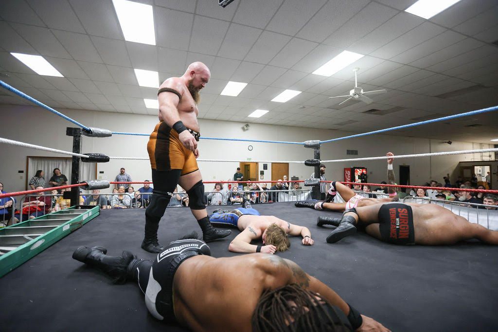 Wrestlers lying on a mat in a gym while a bald wrestler stands over them under bright overhead lights