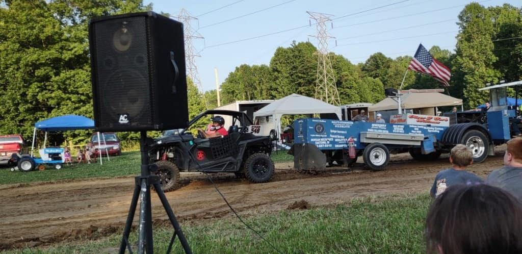 ATV and blue truck towing a trailer at an outdoor event, with a large speaker in the foreground.
