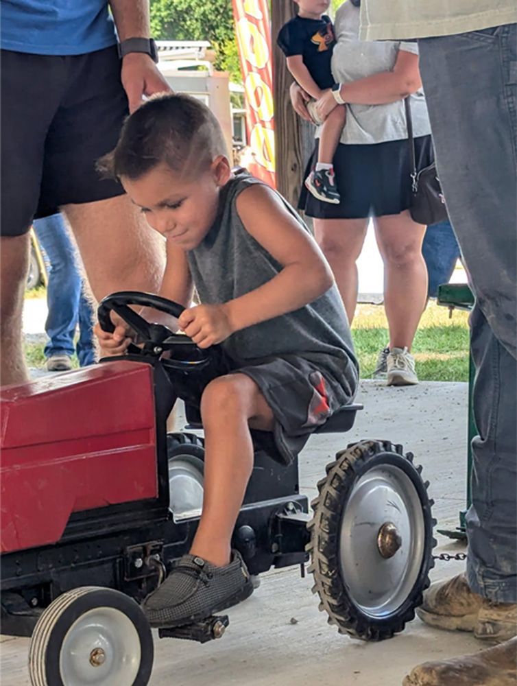 Child driving a small red toy tractor at an outdoor event, surrounded by adults.