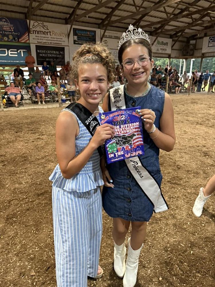 Two girls holding a ribbon and award flyer at a muddy indoor event, smiling in boots and dresses