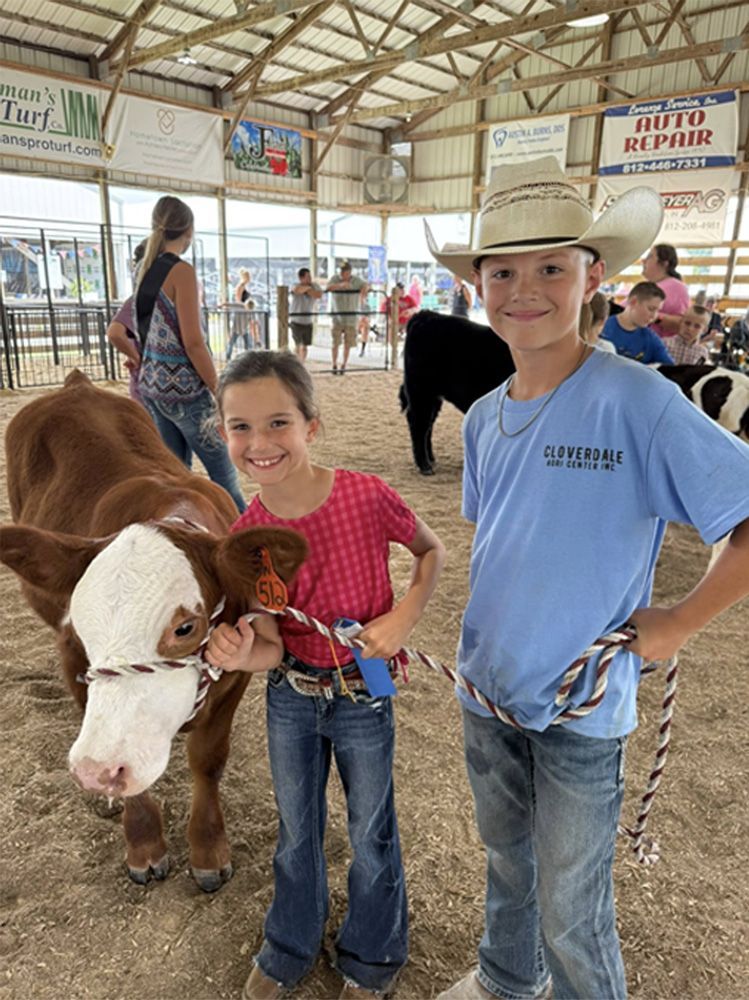 Two children at a livestock fair, smiling beside a calf in a barn with pens and signs in the background
