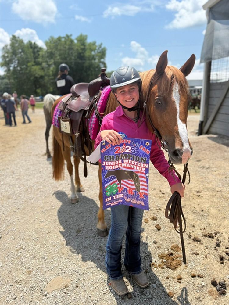 Girl smiling beside a saddled horse on a gravel path, holding the reins at an outdoor event.