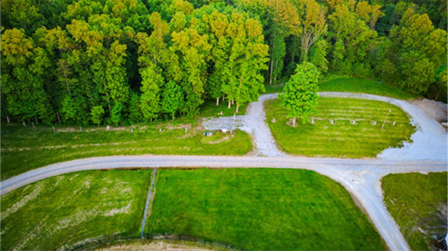 Aerial view of a grassy park with winding paths, open green field, and a dense tree line in the background.