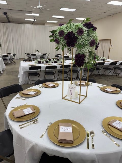 Round banquet table with gold place settings and a tall green-and-purple floral centerpiece in a reception hall