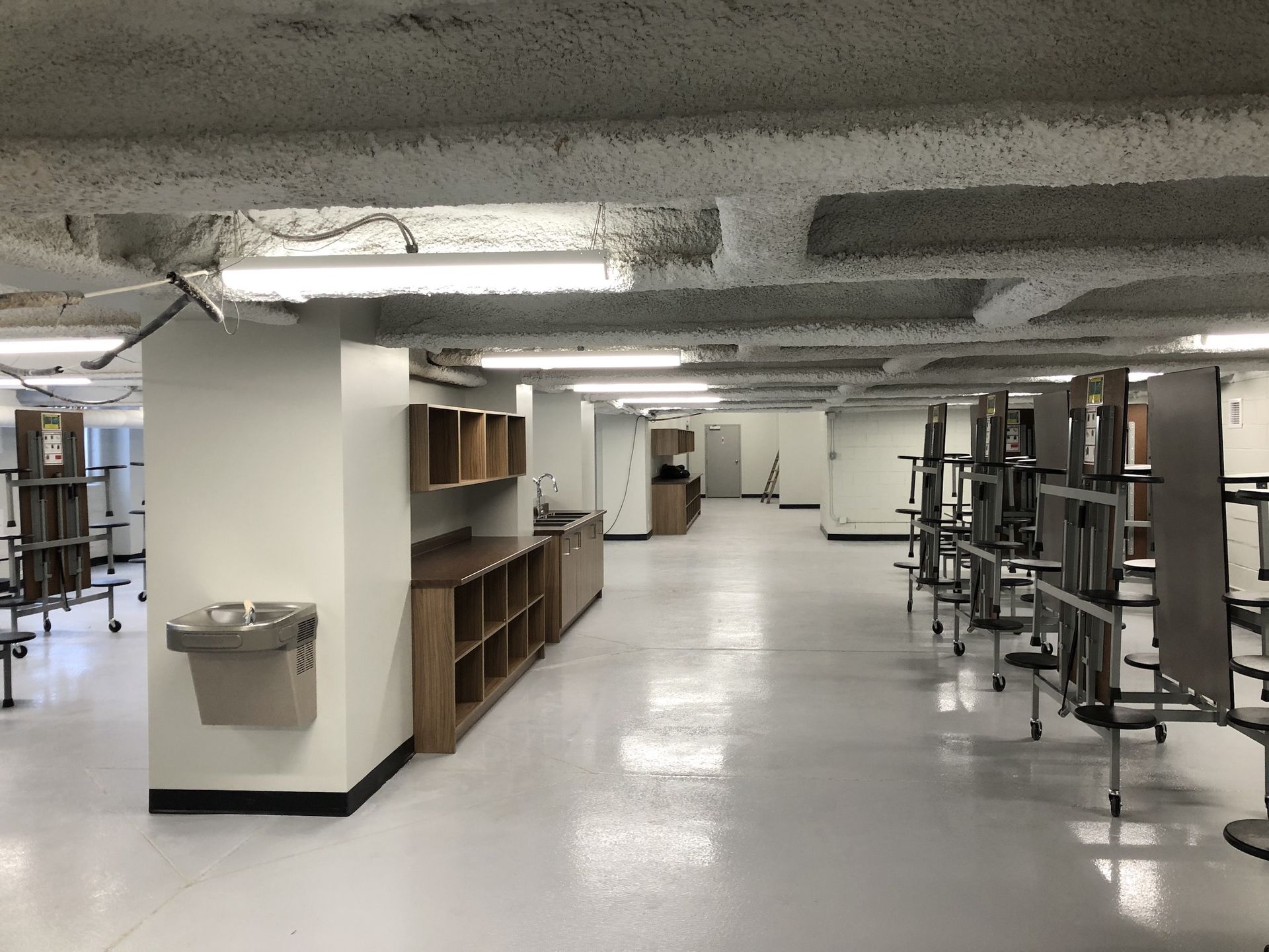 Cafétéria institutionnelle vide avec des tables empilées, des murs blancs et un plafond moucheté.