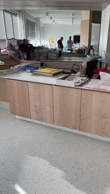 Comptoir avec armoires et objets en bois clair. Deux personnages en arrière-plan. Bureau lumineux et moderne.