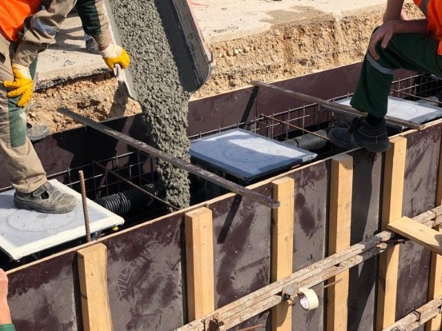 Workers pouring concrete into a wooden formwork. Grey cement flows, seen from a high angle.