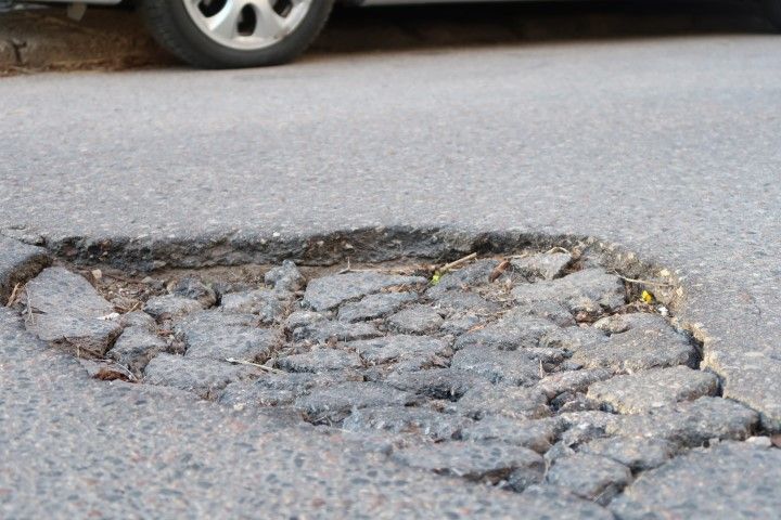 Close-up of a large pothole in asphalt road, near a car's tire. Damaged surface with broken pieces.