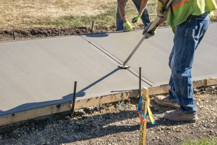 Workers smoothing wet concrete for a sidewalk with hand tools; wood forms in place.