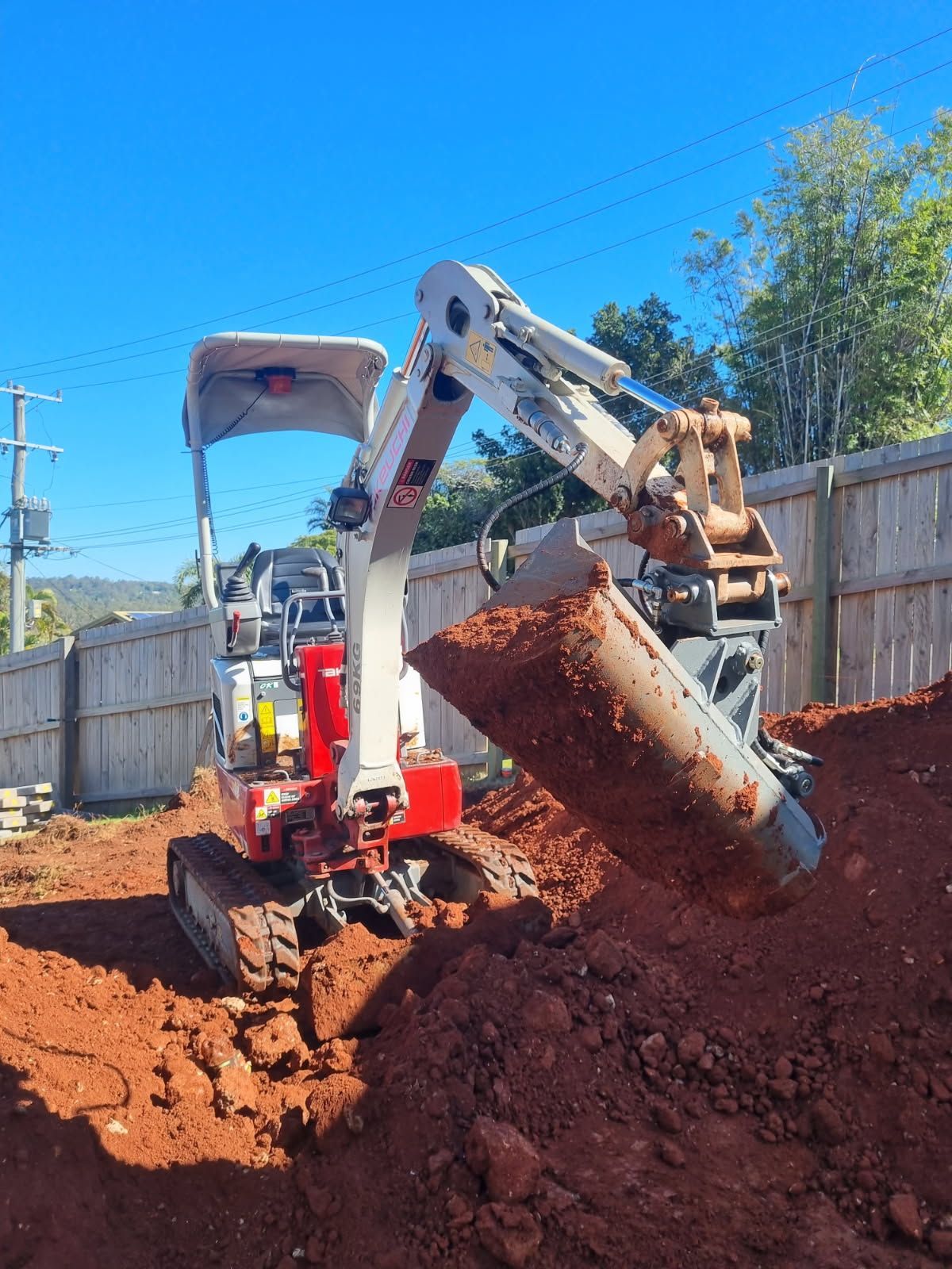 Mini excavator digging into red soil. Blue sky in the background, wooden fence. — Coastline Earthworks in Helensvale, QLD
