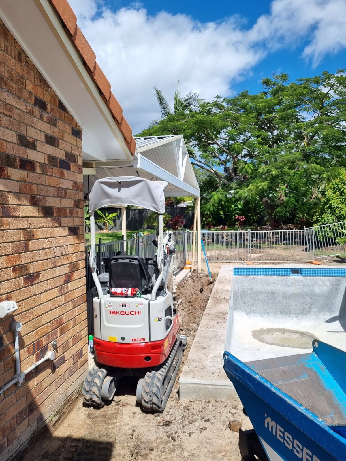 Mini excavator next to a brick wall and a pool, digging a trench. Blue sky with trees in the background — Coastline Earthworks in Helensvale, QLD